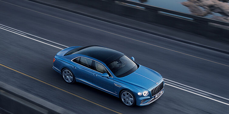 Bentley Mougins New Bentley Flying Spur Azure sedan in Light windsor blue paint by Mulliner, with 22 inch Azure wheels seen from overhead driving dynamically on a country road