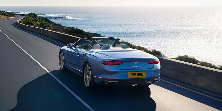 Bentley Mougins New Bentley Continental GTC Azure convertible rear view in Light Windsor blue paint with 22 inch ten spoke wheels driving dynamically on an ocean road in the sunshine