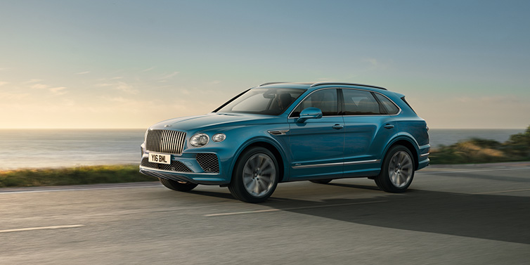 Bentley Mougins Bentley Bentayga Azure SUV in Topaz blue paint driving dynamically by the ocean with 22 inch 10 spoke directional wheels