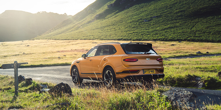 Bentley Mougins Bentley Bentayga S SUV rear three quarter in Sunburst Gold paint on a road surrounded by green hills