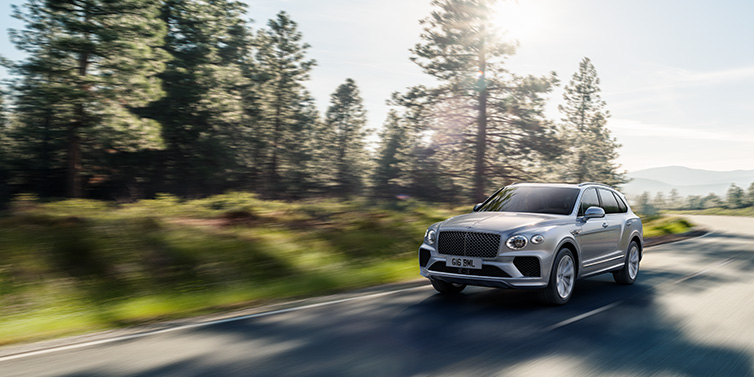 Bentley Mougins Bentley Bentayga SUV front three quarter in Moonbeam silver paint driving dynamically along a tree-lined road