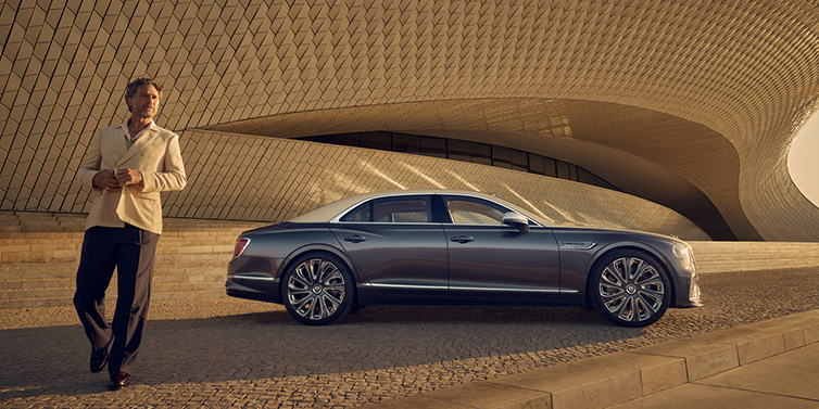 Bentley Mougins Bentley Flying Spur Mulliner sedan in Rose Gold over Magnetic duotone paint parked by a curved wall with person in suit jacket standing on the left